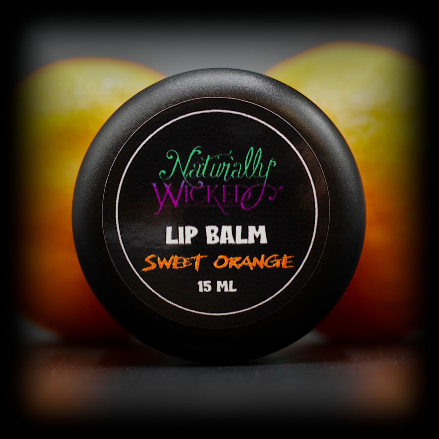 Naturally Wicked Sweet Orange Lip Balm Vibrant Lid In Front Of Two Vivid Orange Fruits