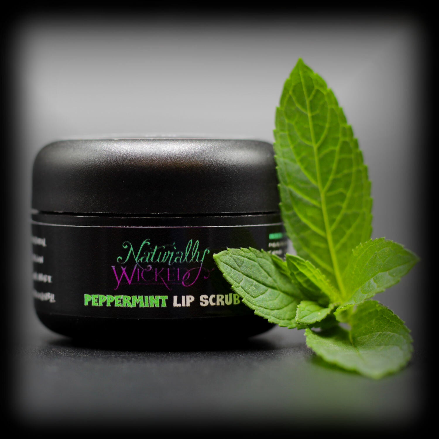 Naturally Wicked Peppermint Lip Scrub In Luxury Black Container Beside Fresh Green Peppermint Leaf