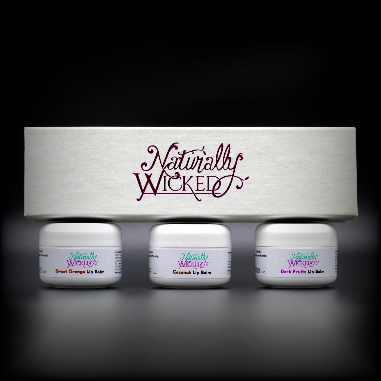 Naturally Wicked Lip Balm Trio Box On Top Of 3 Luxury Fruit Filled White Lip Balm Containers; Coconut, Dark Fruits & Sweet Orange