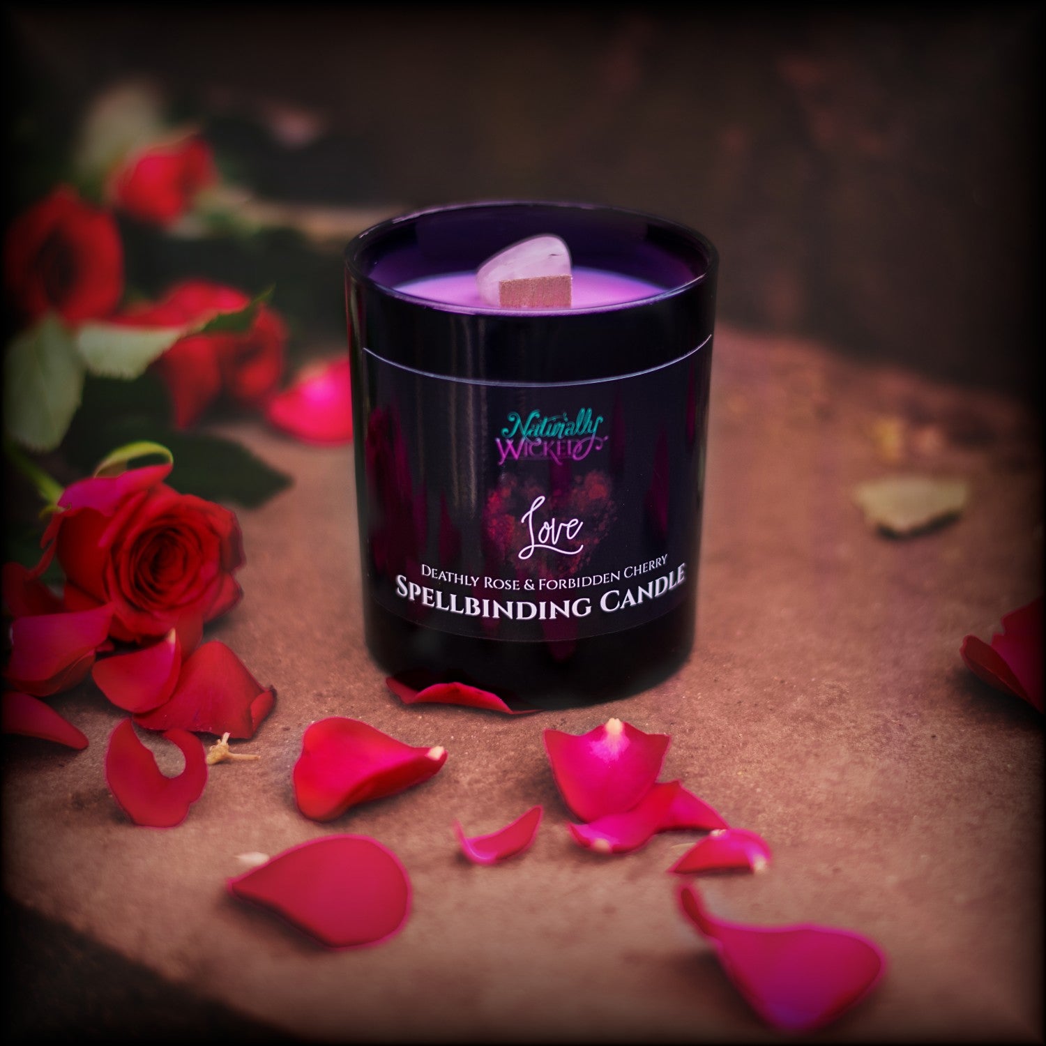 A Scenic View Of The Perfect Spell Candle Surrounded By Red Roses And Red Rose Petals. Naturally Wicked Spellbinding Love Candle Proudly Presents It's Dark Black Gloss Label With A Bright Red Love Heart On The Front. The Candle Features Plant-based Smooth Pink Wax, A Wood Wick And A Beautiful Rose Quartz Crystal.