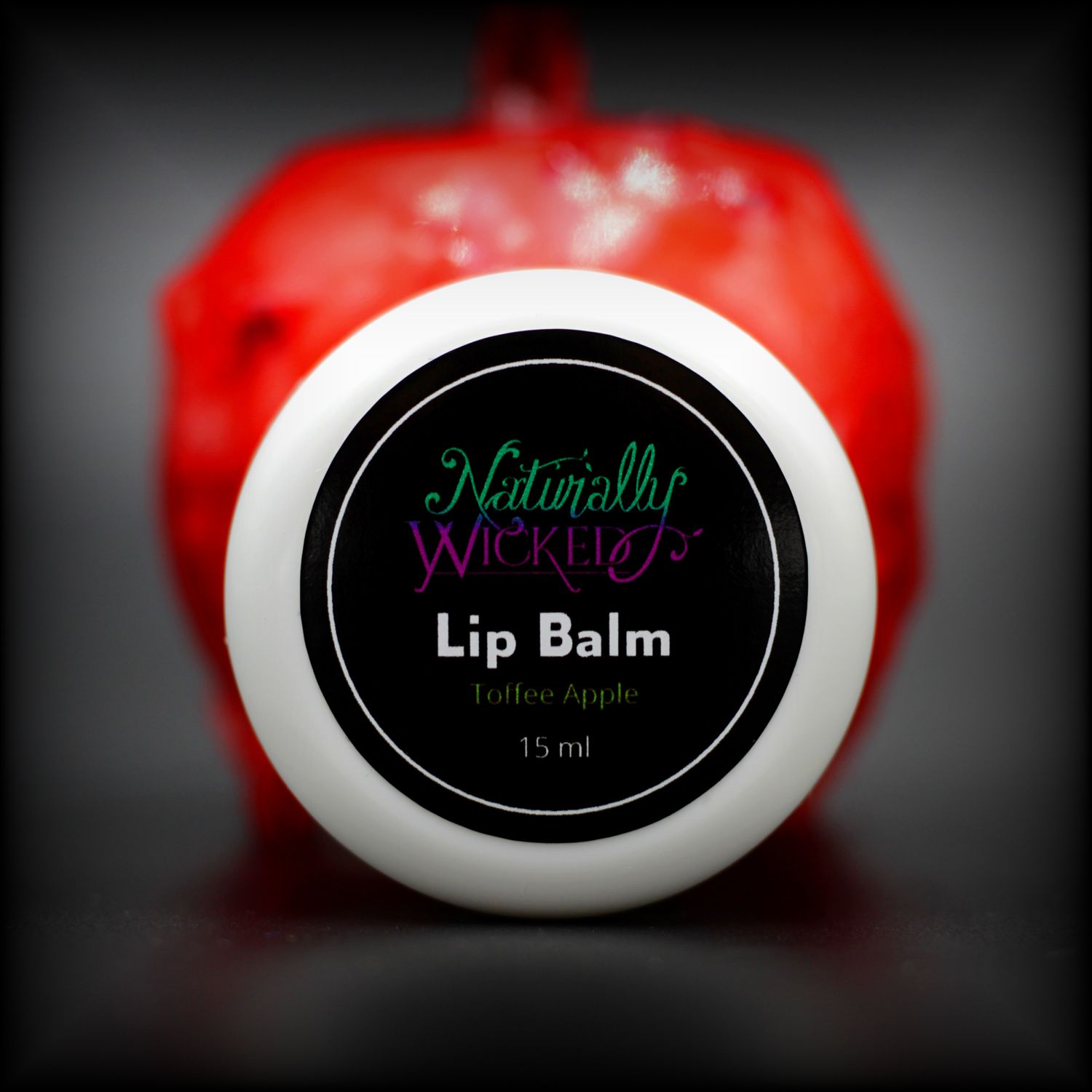 Naturally Wicked Toffee Apple Lip Balm Lid Face In Front Of Bright Red Toffee Apple 