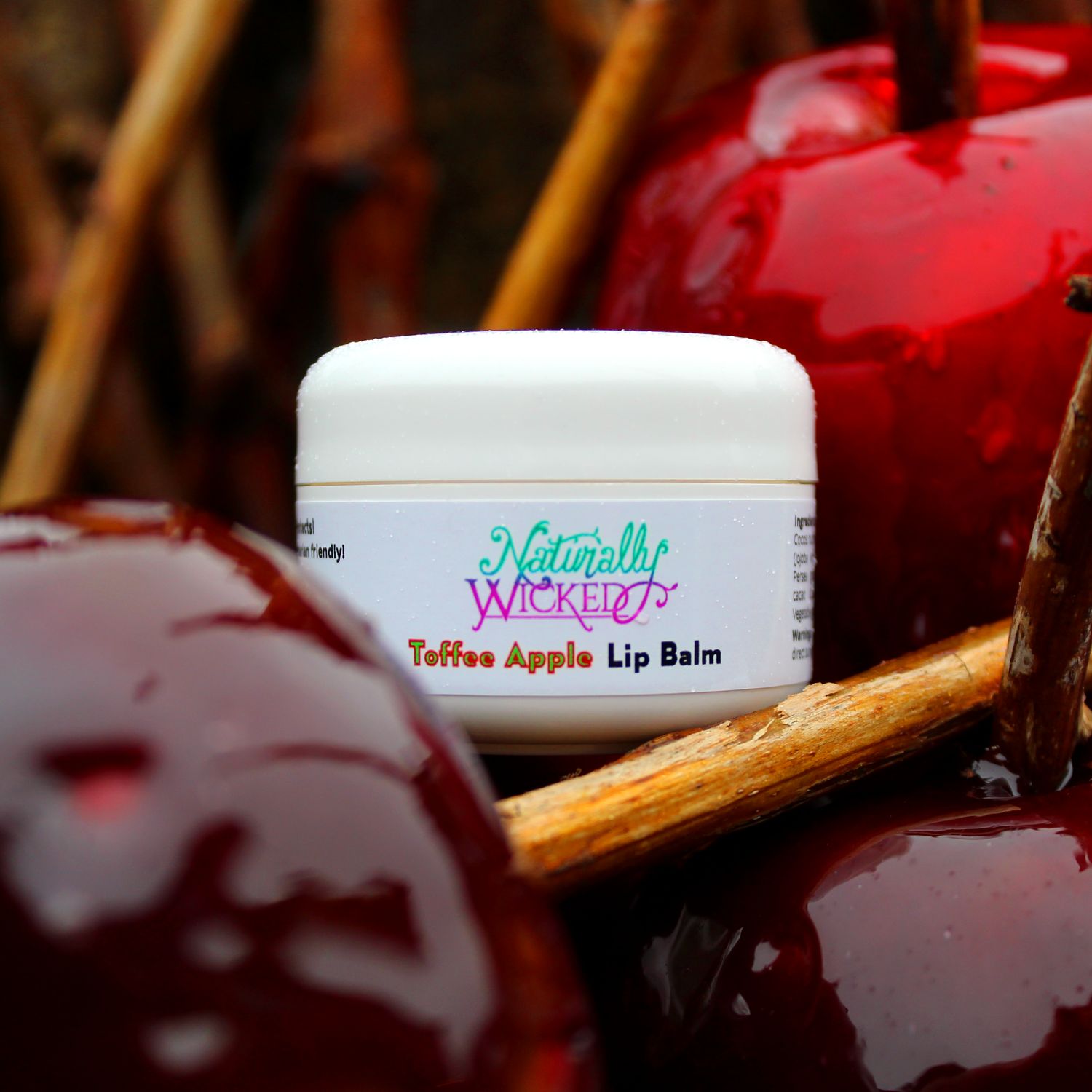 Naturally Wicked Toffee Apple Lip Balm Surrounded By Bright Red & Purple Glossy Toffee Apples & Brown Sticks